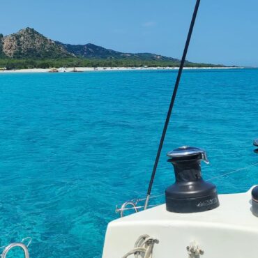 Blick über die Winschen des Katamarans auf das türkisfarbene Wasser und den weißen Sandstrand an der Ostküste Sardiniens.